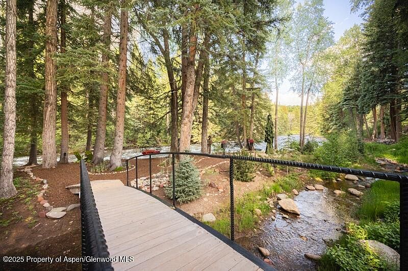 This image showcases a serene yard/garden setting with a wooden bridge spanning a small stream. Lush greenery and mature trees surround the area, creating a tranquil and natural ambiance. The scene suggests a peaceful retreat, ideal for relaxation and enjoying the outdoors.