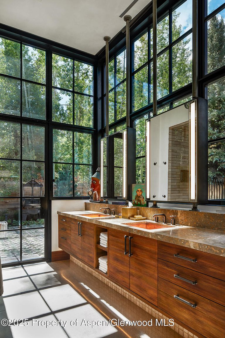 This is a luxurious primary bathroom featuring a double vanity with copper sinks and dark wood cabinetry. Large windows offer a view of the surrounding trees, bringing natural light into the space. The mirrors are framed with integrated lighting, adding a modern touch to the rustic design.