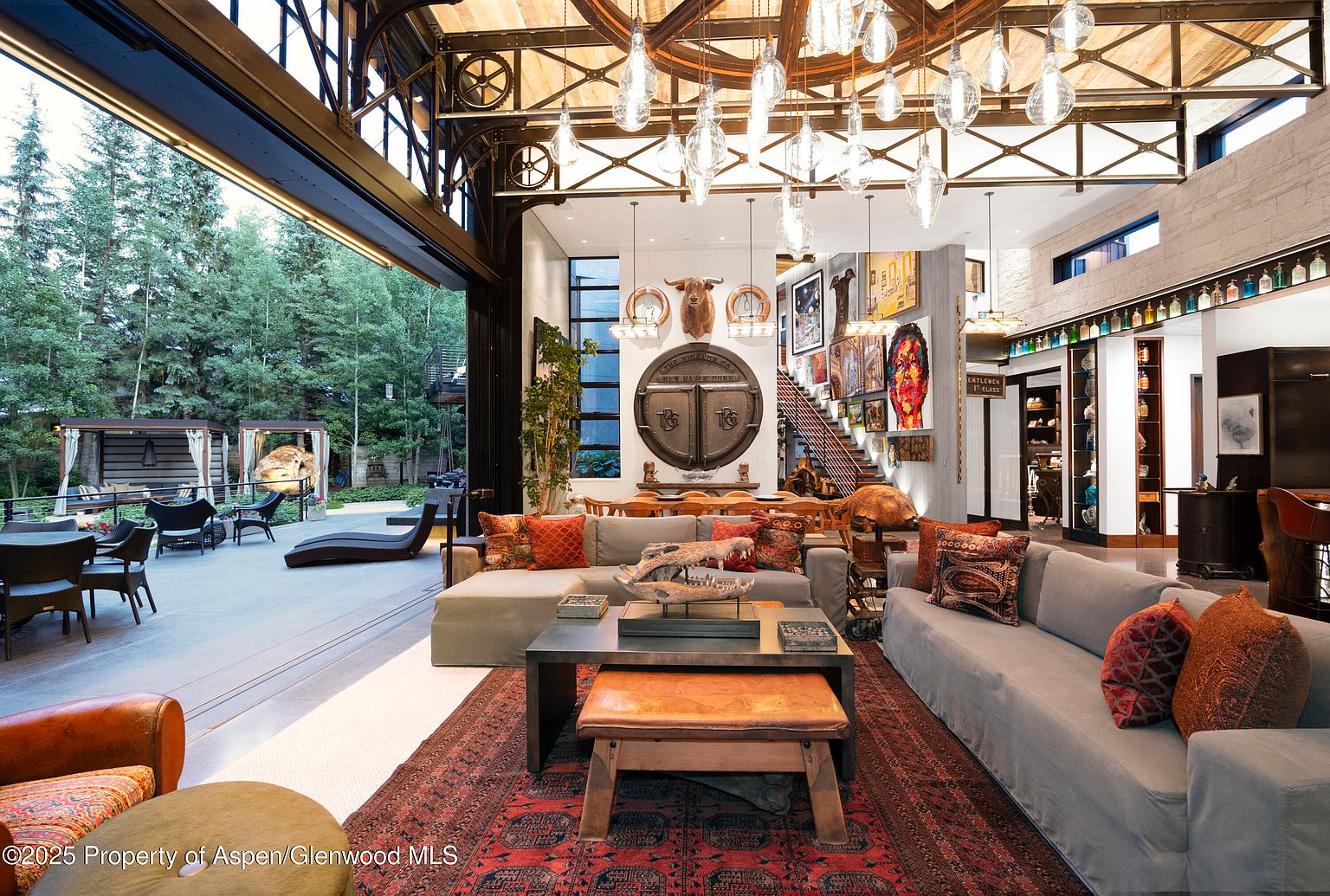 This is a spacious and luxurious living room with an open floor plan that seamlessly connects to an outdoor patio. The interior features a blend of rustic and modern elements, including exposed steel beams, unique light fixtures, and comfortable seating arrangements. The room is decorated with eclectic art and accessories, creating a warm and inviting atmosphere.