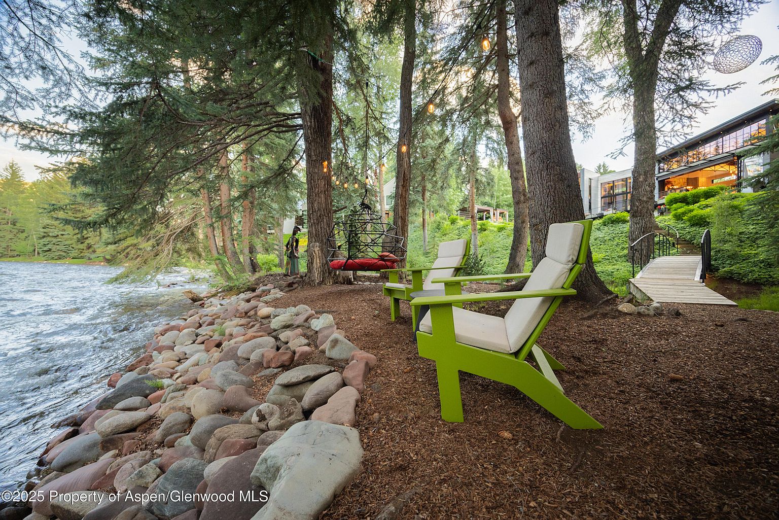This picturesque yard features a riverfront setting with large rocks lining the shore. Two green Adirondack chairs offer a relaxing spot to enjoy the view, while string lights hang between the trees, creating a magical ambiance. A modern house is visible in the background, accessible by a small bridge.