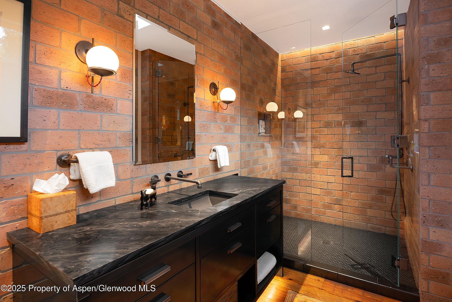 This is a primary bathroom featuring exposed brick walls, a dark vanity with a stone countertop, and a glass-enclosed shower. The vanity has a modern sink and faucet, with globe-shaped sconces providing warm lighting. The shower has a dark tiled floor and a sleek, minimalist design, creating a modern yet rustic aesthetic.