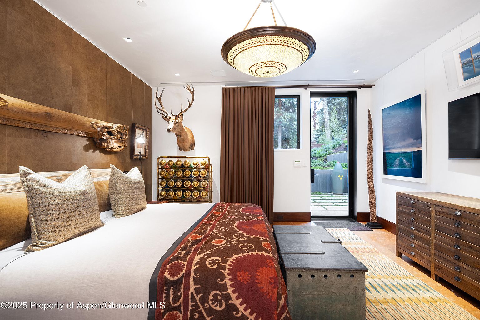 This is a primary bedroom featuring a king-size bed with decorative pillows and a patterned blanket. The room has a rustic-chic design with a wooden headboard, a mounted deer head, and a unique light fixture. A sliding glass door leads to an outdoor area, providing natural light and a connection to the outdoors.