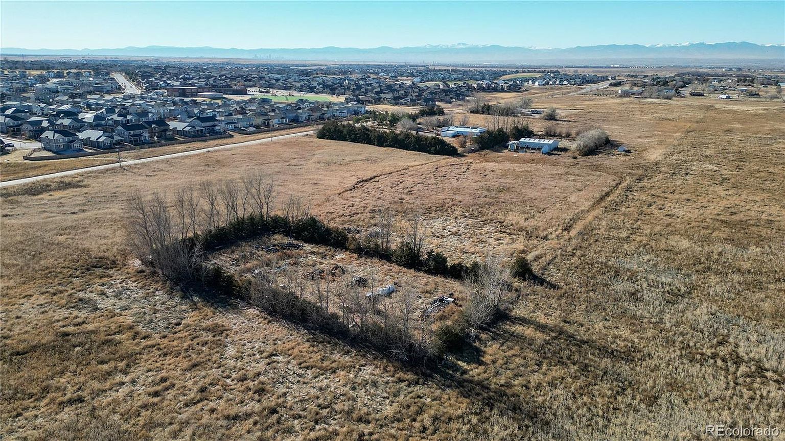 This aerial view showcases a large plot of land with sparse vegetation and a cluster of trees in the foreground. In the distance, a residential area is visible, suggesting the property's proximity to a neighborhood. The landscape is mostly dry and open, offering potential for development or agricultural use.