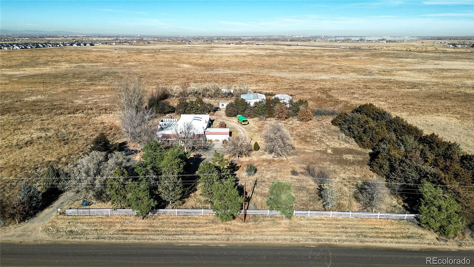 This aerial view showcases a sprawling property featuring a white house with multiple outbuildings, surrounded by a vast, dry landscape. The property is partially enclosed by a white fence, with clusters of trees providing privacy and visual interest. In the distance, a cityscape is visible, adding context to the property's location.