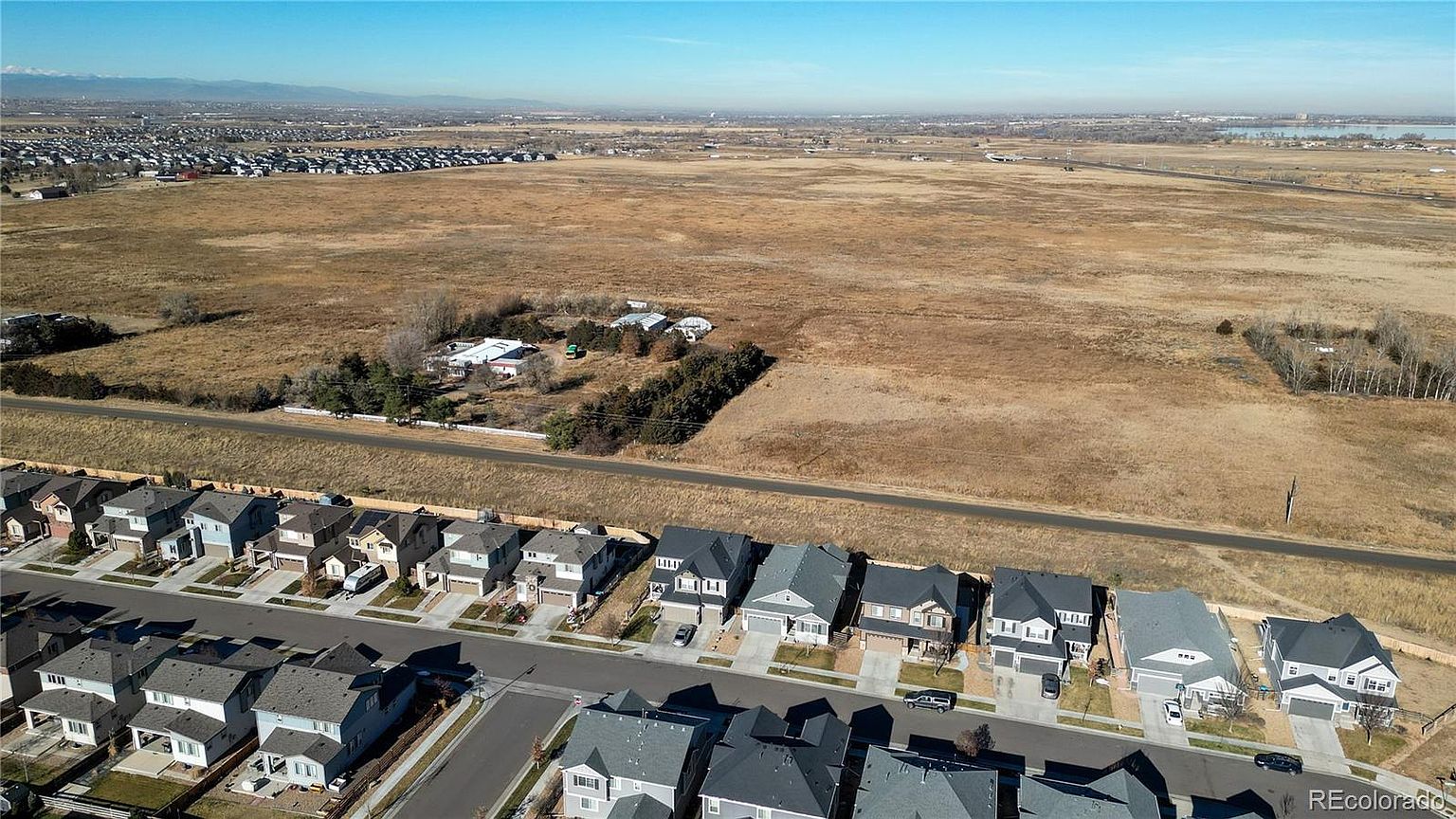This aerial shot showcases a residential neighborhood bordering a vast open field. The houses are neatly arranged with well-maintained lawns and driveways, presenting a clean and orderly appearance. The expansive field provides a sense of openness and potential for future development or recreational space, adding to the property's appeal.