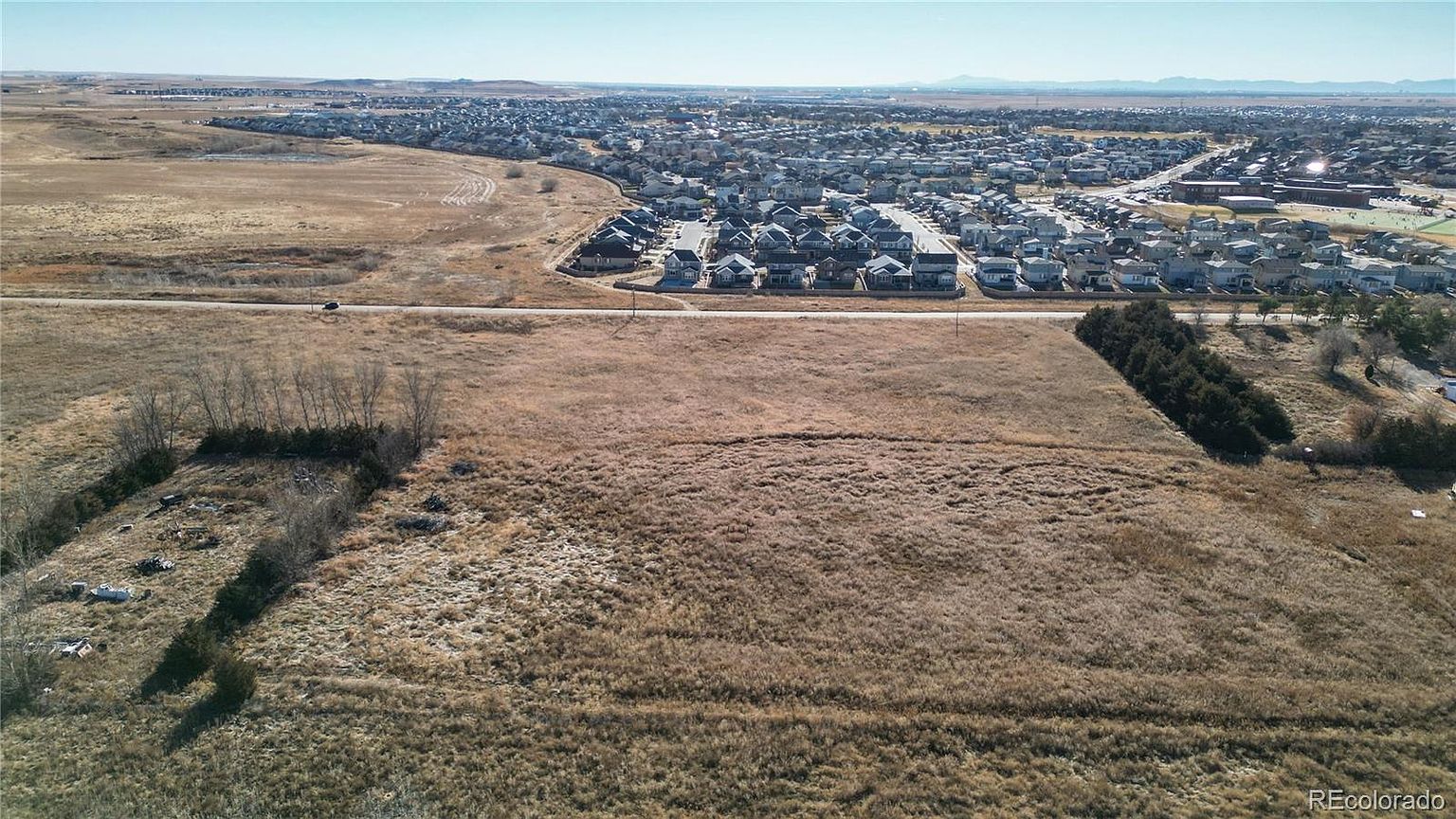 This aerial view showcases a large plot of land with dry, golden grass, bordered by trees and a road. In the background, a residential neighborhood stretches out, offering a sense of community and proximity to amenities. The image provides a comprehensive view of the property's potential and its surroundings.