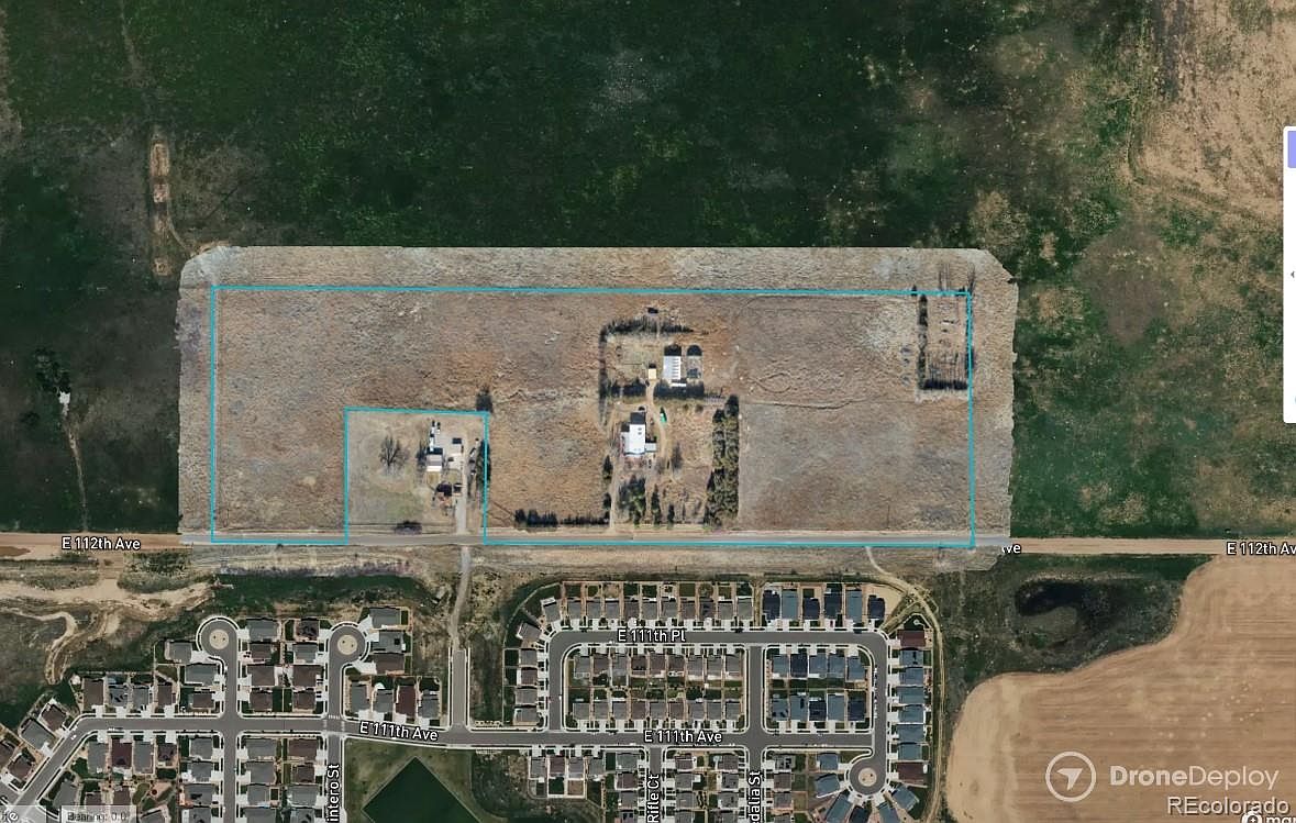 This aerial view showcases a large property with a mix of open land, residential buildings, and surrounding neighborhood. The property is bordered by a road and features a couple of houses with outbuildings, while the adjacent area consists of neatly planned residential developments. The overall impression is one of a spacious property with potential for various uses, contrasted by the organized suburban landscape nearby.
