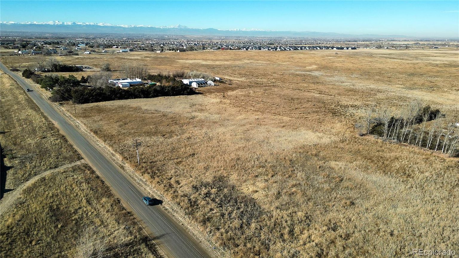 This aerial view showcases a vast, dry landscape with a road cutting through the scene. A cluster of buildings is nestled among trees in the mid-ground, while a distant town and snow-capped mountains provide a scenic backdrop. The perspective emphasizes the property's expansive surroundings and potential for development.