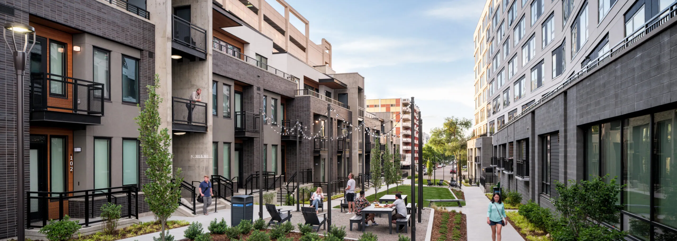 This image showcases an attractive outdoor space between modern apartment buildings. There is a landscaped area with seating, walking paths, and string lighting, creating a community gathering spot. The well-maintained landscaping and inviting atmosphere add to the property's appeal.