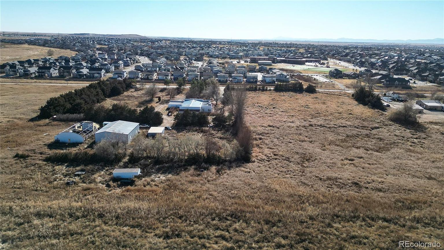This aerial view showcases a large plot of land with several outbuildings, including a metal barn and a smaller structure, surrounded by dry, golden-brown grass. In the background, a residential neighborhood stretches towards the horizon under a clear blue sky. The image provides a sense of the property's size and its proximity to a developed area.
