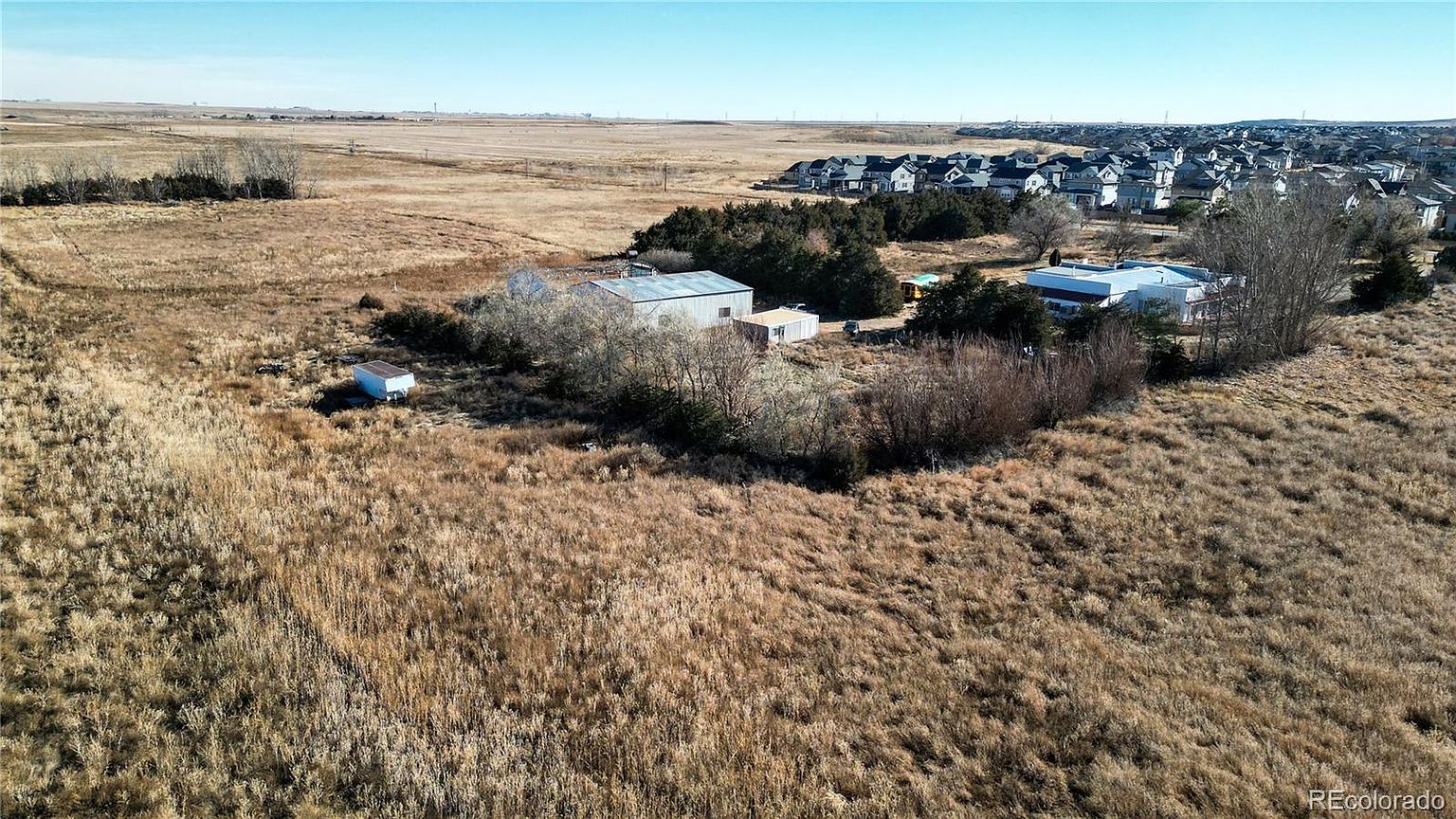 This aerial view showcases a large, undeveloped lot with dry, golden grass and scattered trees. Several buildings are situated on the property, including a metal-roofed structure and a smaller outbuilding. In the background, a residential neighborhood is visible, suggesting potential for future development or expansion.