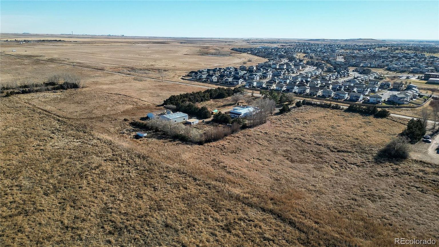 This aerial view showcases a property situated in a transitional landscape, blending rural and suburban elements. The property includes several buildings surrounded by open fields, with a developing residential area visible in the background. The image highlights the property's spacious setting and proximity to both natural and community amenities.