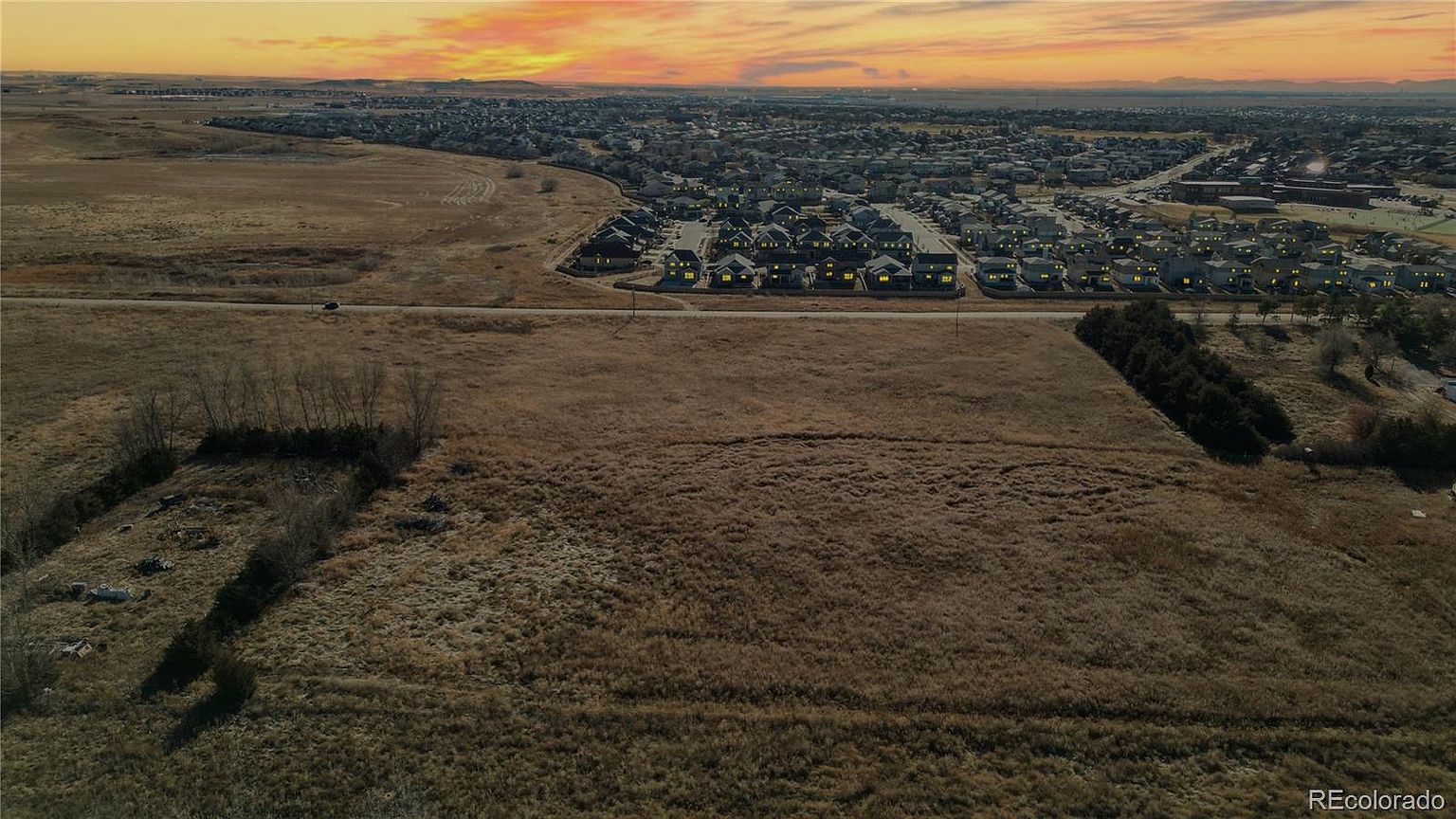 This aerial view showcases a vast, undeveloped field in the foreground, transitioning to a residential neighborhood in the background. The houses are neatly arranged with visible lighting, suggesting a well-planned community. The sky displays a warm sunset, adding a touch of serenity to the landscape and highlighting the potential for future development or the peaceful setting of the existing homes.