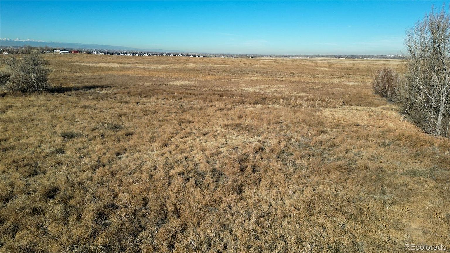 This aerial view showcases a vast, undeveloped land parcel under a clear blue sky. The landscape is predominantly covered in dry, golden-brown grasses, suggesting a field or prairie environment. In the distance, a faint line of buildings and possibly mountains can be seen, indicating the proximity to a town or city.