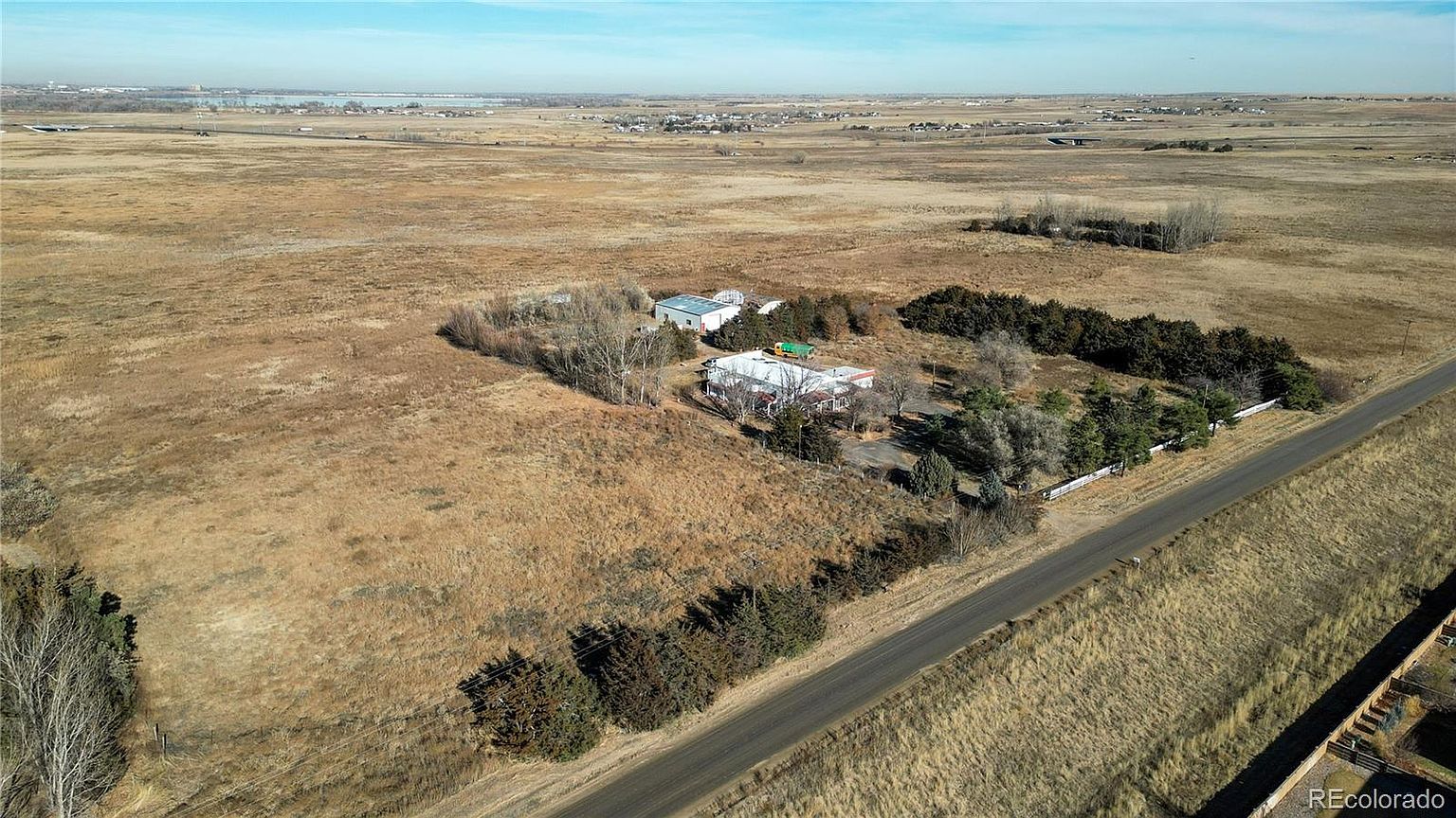 This aerial shot showcases a property featuring a main house and a secondary building, surrounded by a mix of trees and open, grassy land. A road runs along the edge of the property, providing access. The landscape is predominantly dry and rural, giving a sense of seclusion and space.