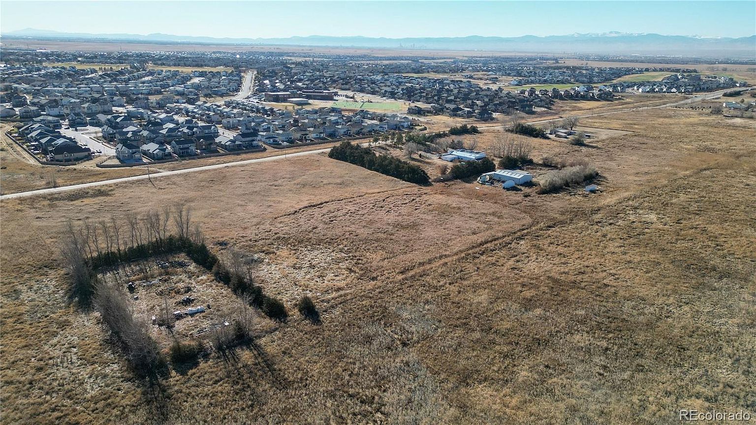 This aerial view showcases a large plot of land with a mix of dry grass and some trees, situated near a residential area. Several buildings are visible on the property, and in the distance, a sprawling neighborhood extends towards the horizon, with mountains faintly visible in the background. The perspective gives a sense of the property's size and its proximity to both nature and community.