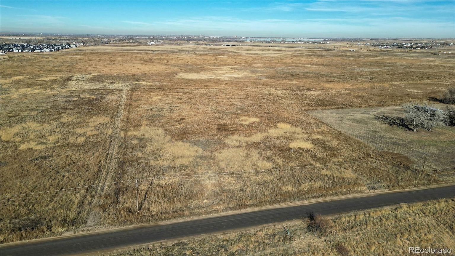 This aerial view showcases a vast expanse of land, primarily consisting of dry grassland with patches of varying textures and colors. A narrow road cuts across the foreground, leading towards a distant horizon where a residential area is visible. The sky is clear and blue, providing a sense of openness and potential for development.