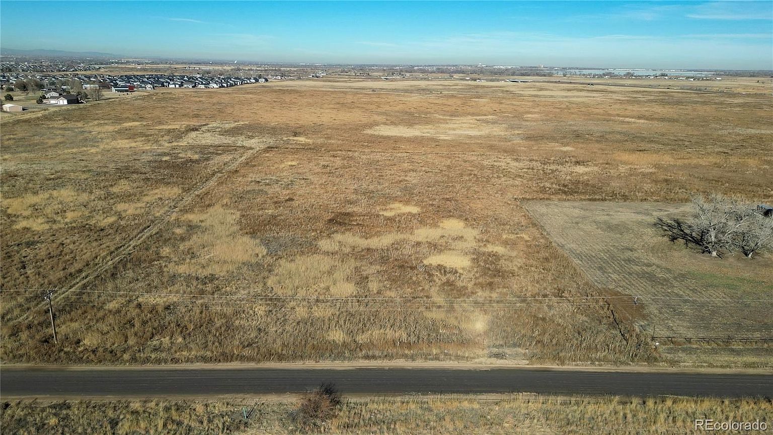This aerial view showcases a vast expanse of land, primarily consisting of dry grassland with varying textures and tones. A road runs along the lower edge of the frame, and in the distance, a residential area is visible, suggesting proximity to a town or city. The overall impression is one of open space and potential development opportunities.