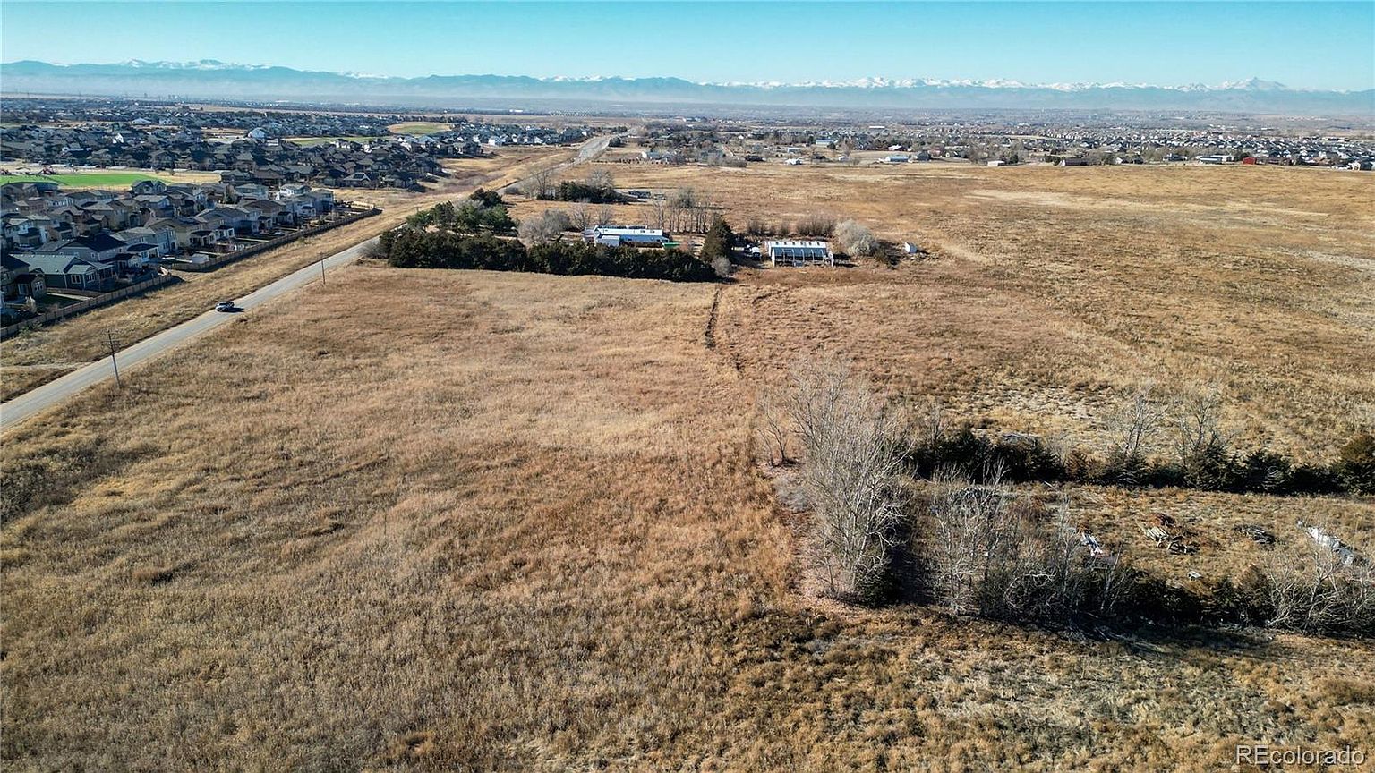 This aerial view showcases a vast, undeveloped plot of land with dry, golden grasses, bordered by a residential area on one side and distant mountains on the horizon. Several buildings are visible in the middle ground, surrounded by trees, suggesting potential for development or agricultural use. The image conveys a sense of spaciousness and opportunity.