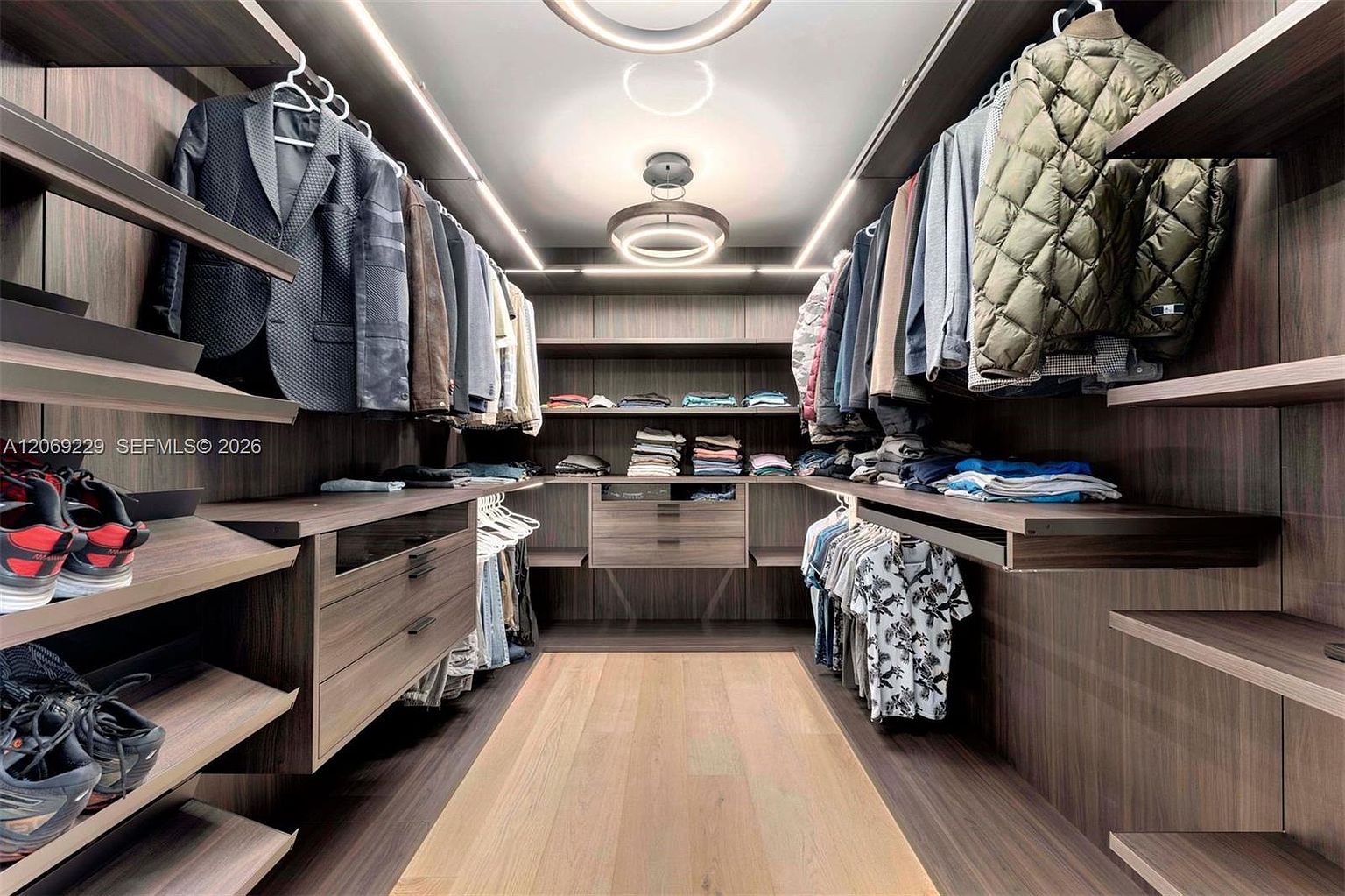 This spacious, modern walk-in closet features dark wood-toned cabinetry and shelving arranged in a symmetrical layout. The space is illuminated by integrated LED lighting strips and a contemporary circular ceiling fixture, creating a sophisticated and organized atmosphere. The perspective is a centered, eye-level shot looking down the aisle, highlighting the ample storage for hanging clothes, folded garments, and shoes.