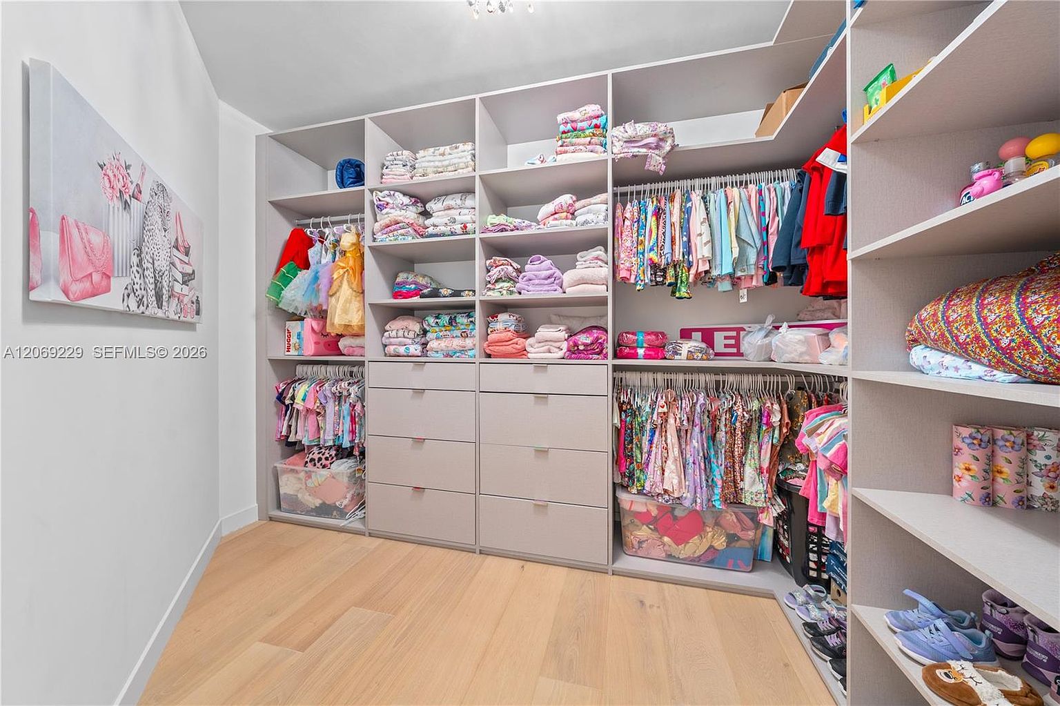 This organized walk-in closet features extensive custom shelving and drawer storage in a light-toned finish. The space is filled with children's clothing, accessories, and toys, showcasing ample storage capacity. A large piece of wall art adds a decorative touch to the room, which is finished with light wood-style flooring.