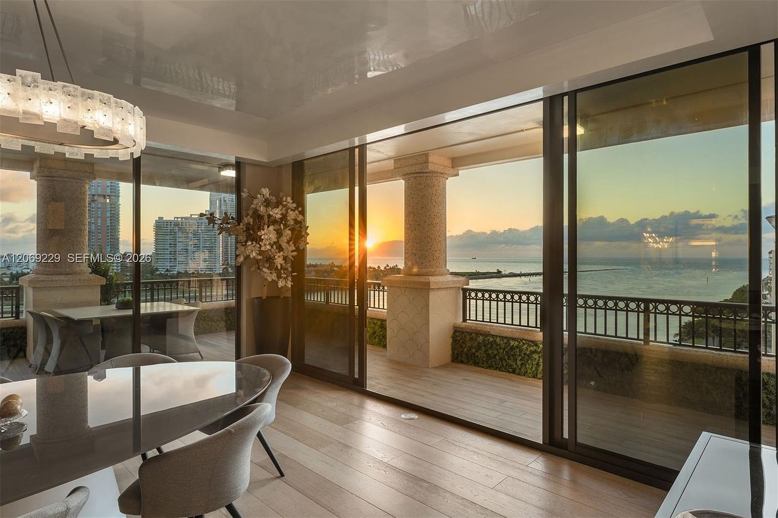 This elegant dining area features a sleek, circular glass-topped table surrounded by modern, light-toned upholstered chairs. The space is defined by expansive, floor-to-ceiling sliding glass doors that open onto a covered balcony, framing a breathtaking sunset view over the ocean and distant city skyline. A sophisticated, multi-tiered crystal chandelier hangs above, adding a touch of luxury to the room's warm, inviting atmosphere.