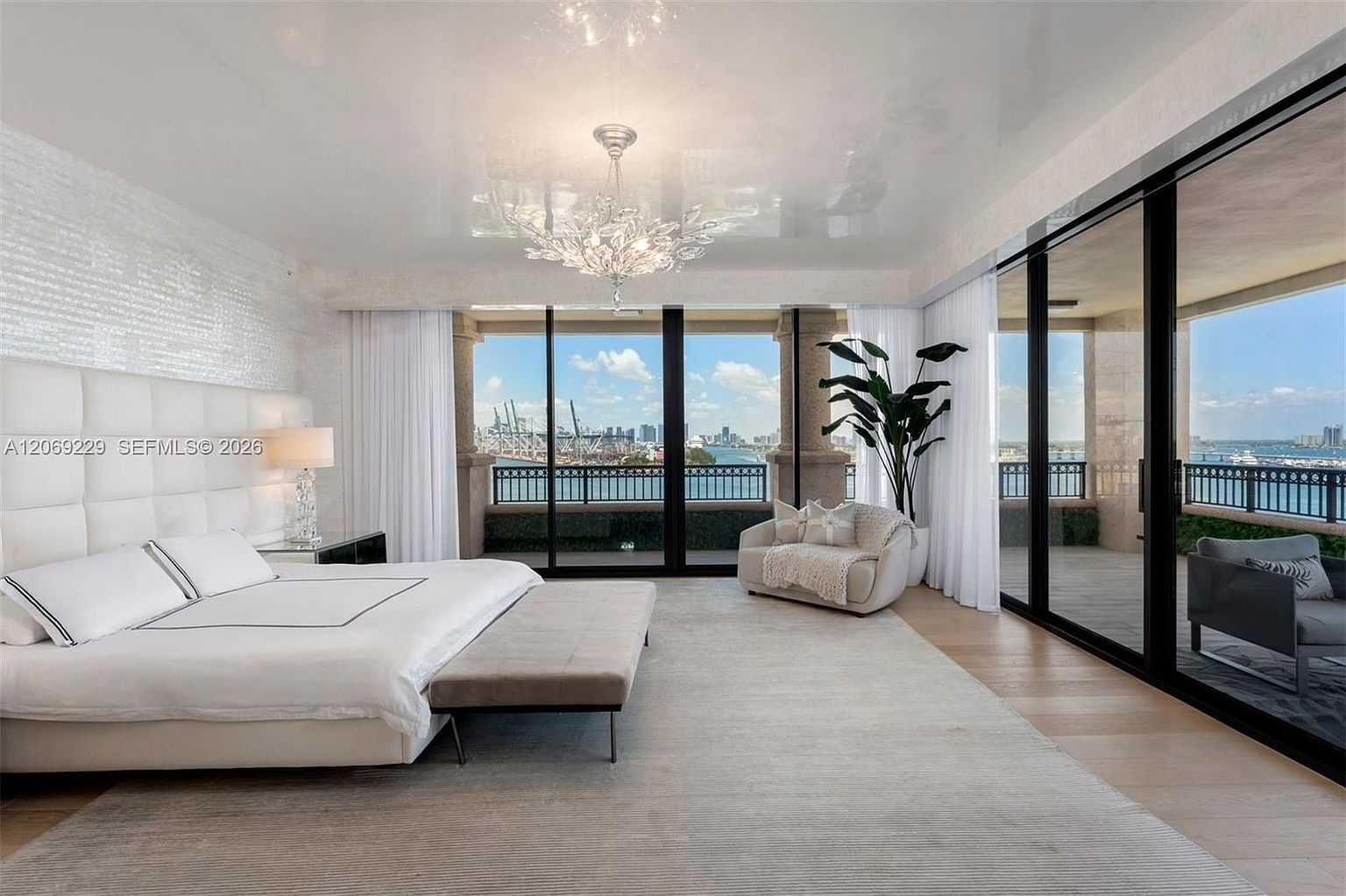 This expansive primary bedroom features a minimalist aesthetic with a large, tufted white headboard and a neutral color palette. The room is defined by floor-to-ceiling windows that provide panoramic views of the waterfront and city skyline, leading out to a private balcony. A sleek chandelier hangs from the reflective ceiling, while a plush area rug anchors the space, creating a serene and luxurious atmosphere.