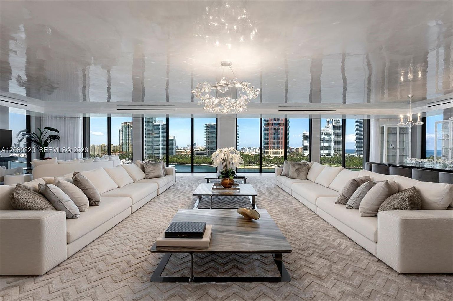 This expansive, symmetrical living room features a high-gloss reflective ceiling and two large, cream-colored sectional sofas facing each other. A pair of low-profile, wood-topped coffee tables sits on a textured, chevron-patterned rug, while floor-to-ceiling windows provide a panoramic view of the city skyline and waterfront. The space is illuminated by a sculptural, branch-like chandelier, creating a sophisticated and airy atmosphere.