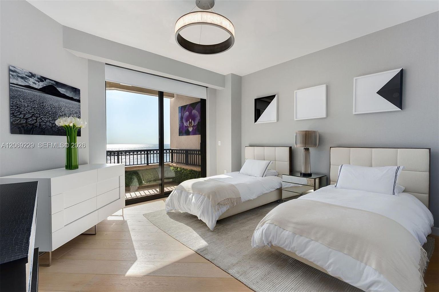 This bright and airy guest bedroom features two twin beds with neutral bedding, positioned on a light-toned area rug over wood flooring. A large sliding glass door provides access to a private balcony with ocean views, allowing ample natural light to fill the room. The space is decorated with minimalist wall art and a modern circular pendant light, creating a clean and contemporary atmosphere.
