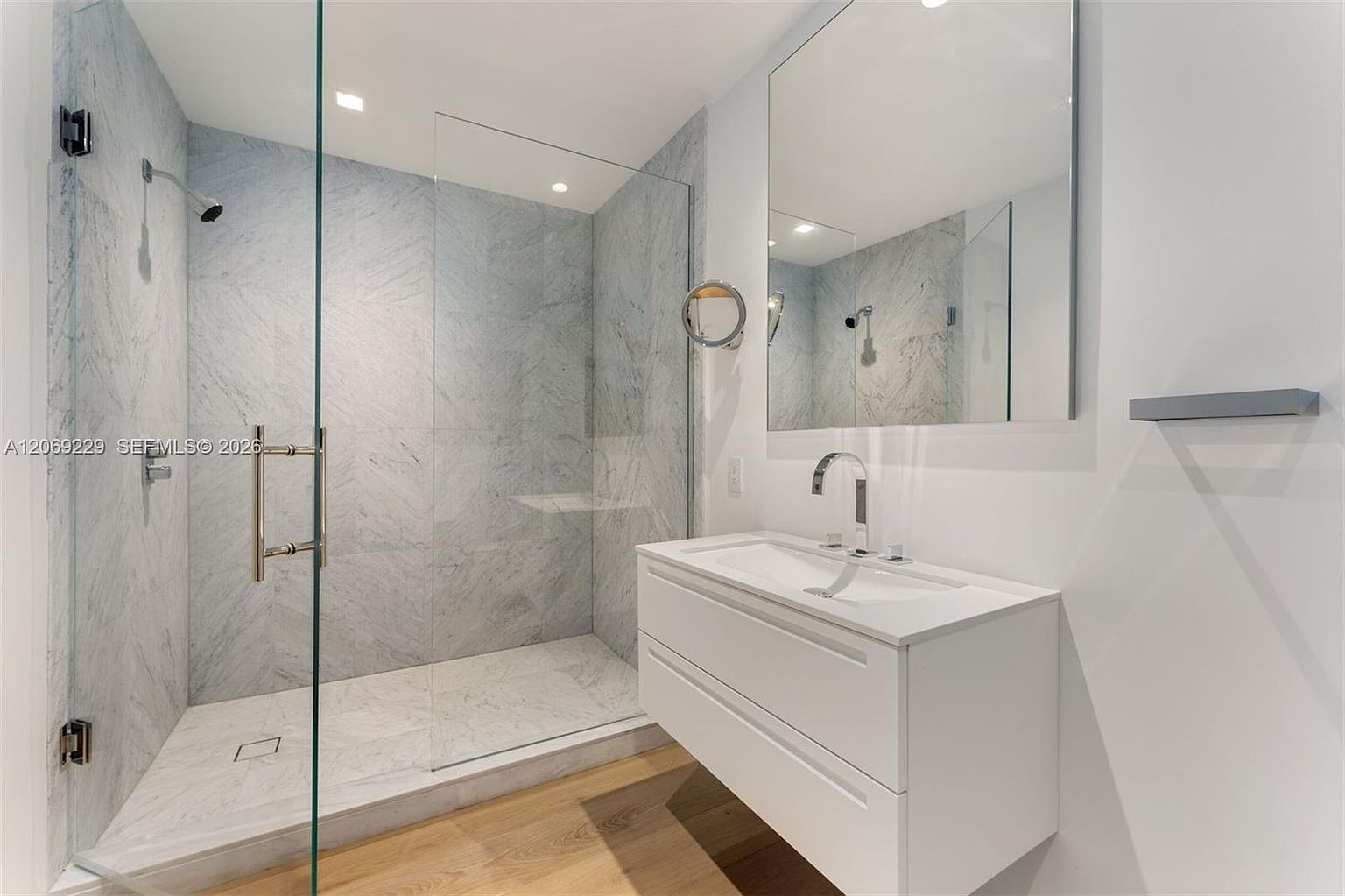 This modern bathroom features a walk-in shower enclosed by glass panels, showcasing light-toned stone wall tiles and a built-in bench. A sleek, wall-mounted white vanity with a single basin sits beneath a large, frameless mirror, complemented by light wood-toned flooring. The space is bright and minimalist, emphasizing clean lines and a contemporary aesthetic.
