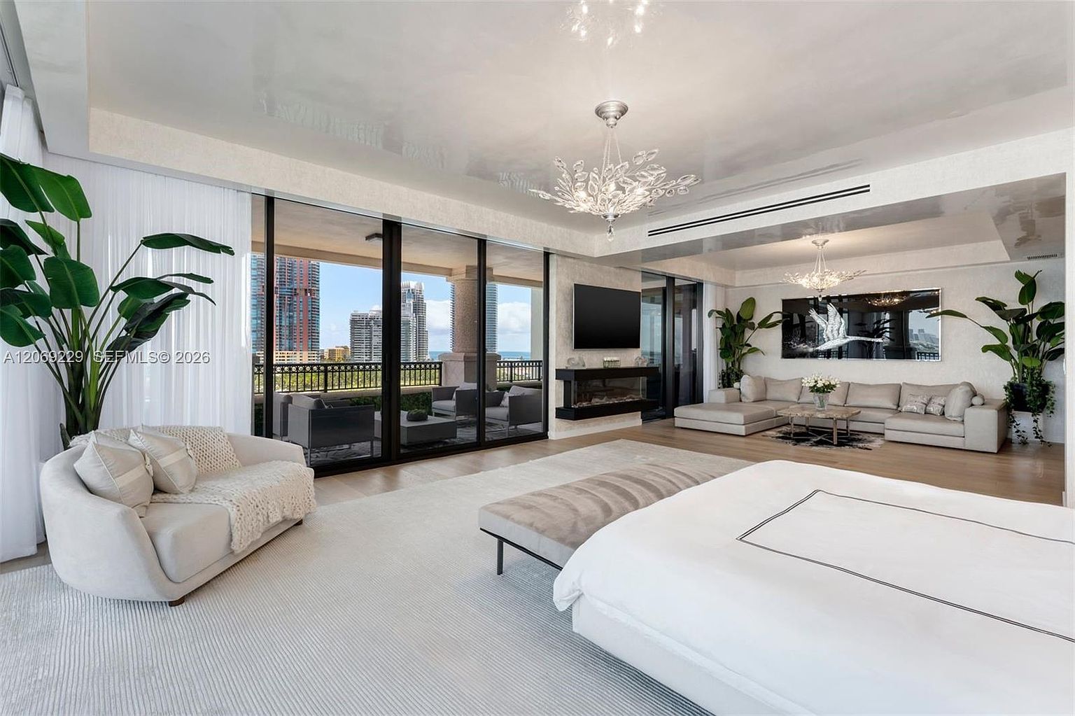 This expansive primary bedroom features a contemporary design with a neutral color palette and a seamless transition to a private balcony through large glass sliding doors. The room is anchored by a plush bed and a bench, complemented by a separate seating area with a sectional sofa and a wall-mounted television above a linear fireplace. The space is illuminated by elegant crystal chandeliers and accented with large potted plants, creating a sophisticated and airy atmosphere.