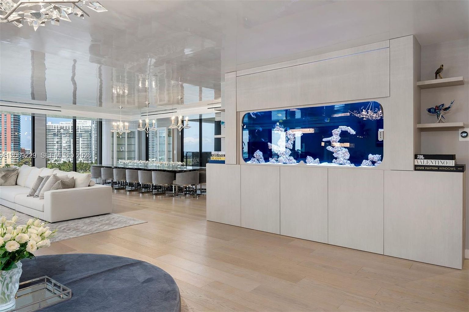 This sophisticated open-concept living space features a large, built-in aquarium integrated into a light-toned cabinetry wall, serving as a striking focal point. To the left, a plush white sofa sits on a patterned rug, while the background reveals a formal dining area with a long table and numerous chairs. The room is characterized by a high-gloss reflective ceiling, light wood flooring, and expansive windows that provide a panoramic view of the city skyline.
