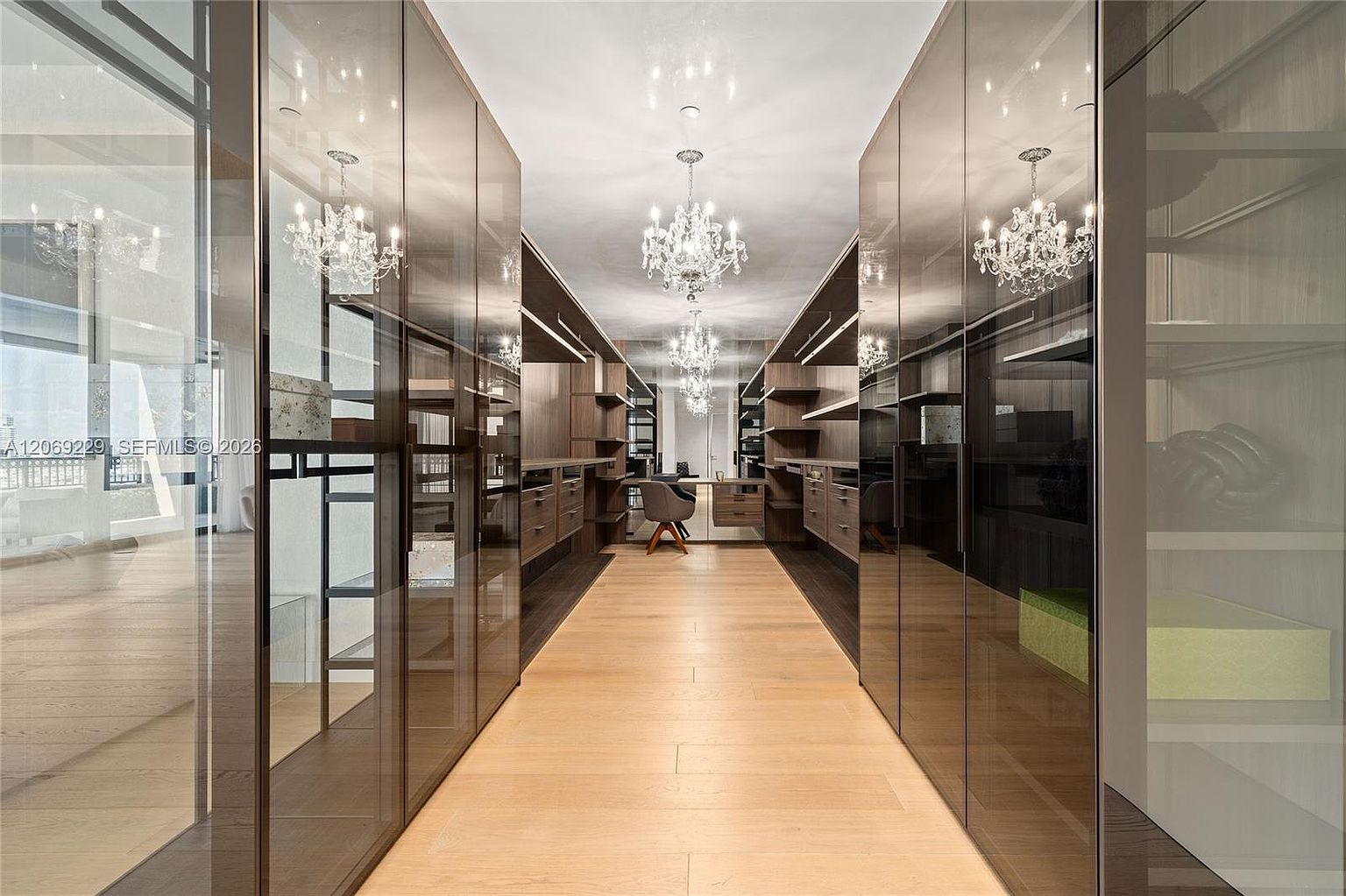 This luxurious walk-in closet features a symmetrical layout with dark wood cabinetry, glass-fronted doors, and integrated shelving. The space is illuminated by two ornate crystal chandeliers, while light wood flooring runs throughout the center aisle. At the far end, a small desk and chair create a functional workspace, reflecting the sophisticated and organized design of the room.