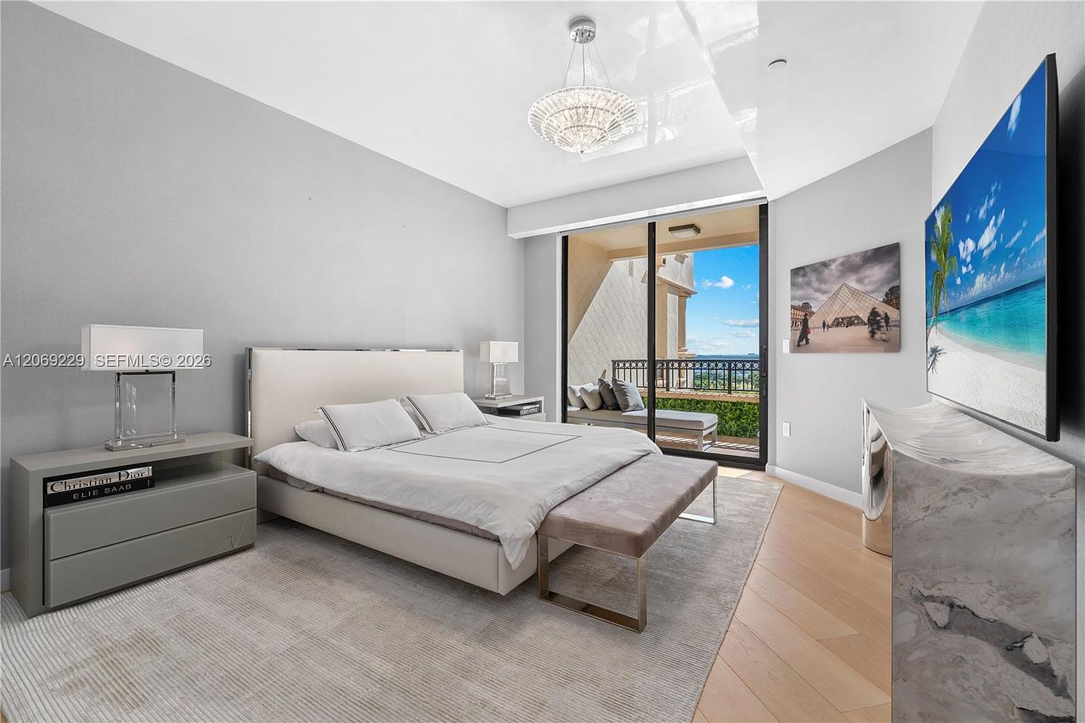This primary bedroom features a contemporary design with a neutral color palette, light wood flooring, and a large bed centered against a soft gray wall. A sleek, light-colored nightstand holds a modern lamp, while a bench sits at the foot of the bed, and a large television is mounted on the wall opposite. Large sliding glass doors open to a private balcony, providing an expansive view of the outdoors and filling the room with natural light.