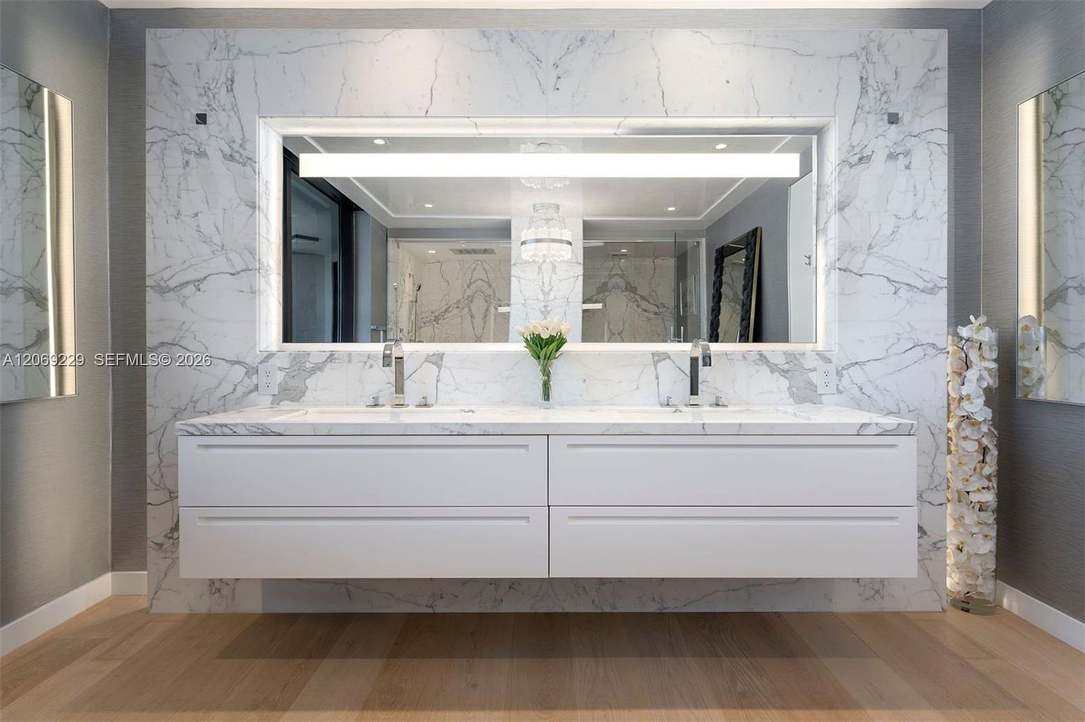 This modern primary bathroom features a floating double vanity with a sleek white finish, set against a striking wall of large-format stone slabs with dramatic veining. A wide, illuminated mirror spans the width of the vanity, flanked by two vertical mirrors on the side walls, while light-toned wood flooring adds warmth to the space. The composition is symmetrical and clean, highlighted by a decorative floral arrangement and a tall, illuminated cylindrical sculpture.