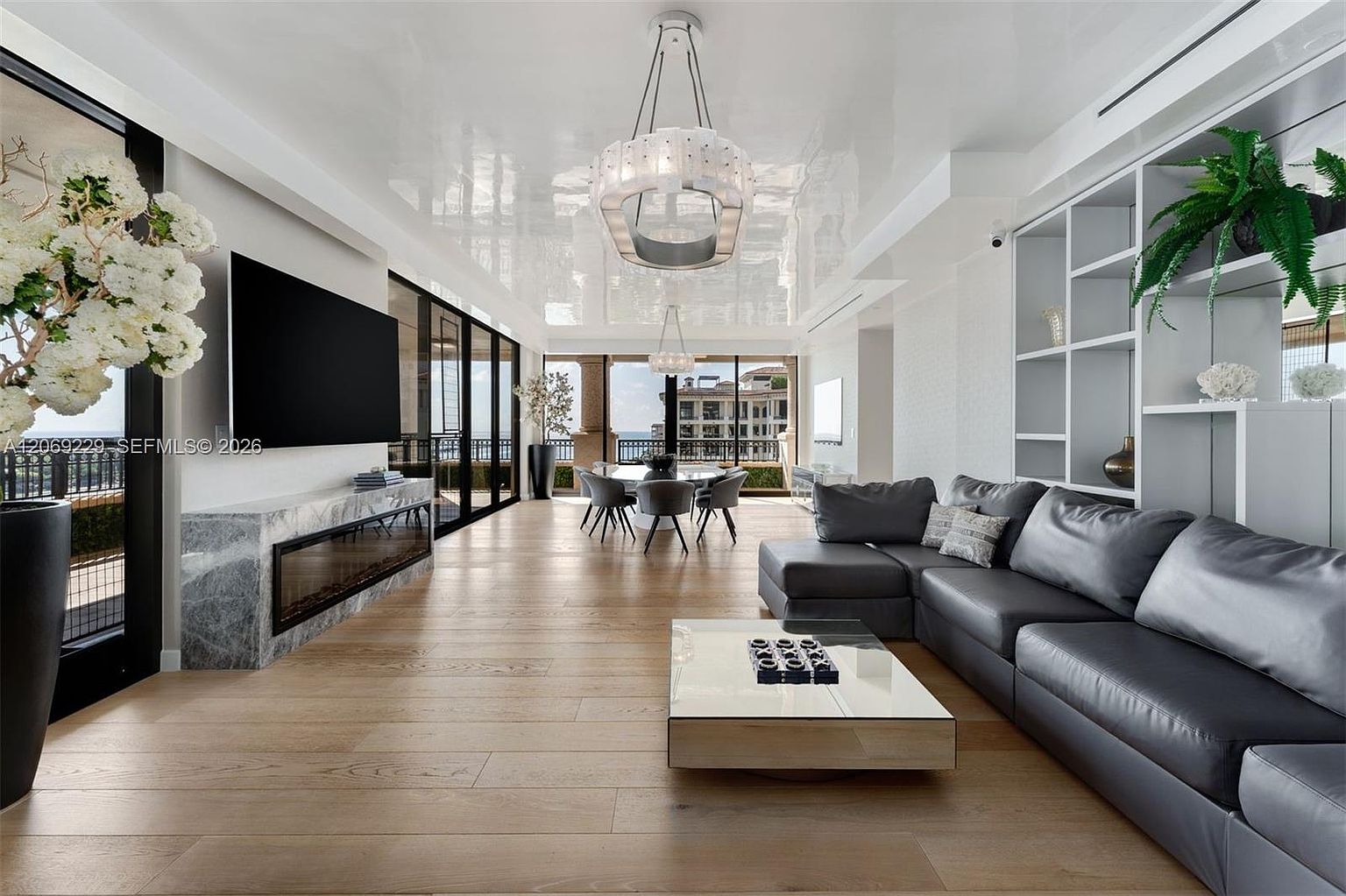 This expansive, open-concept living area features a sleek, dark leather sectional sofa and a low-profile coffee table set against a backdrop of light wood flooring. A linear fireplace is integrated into a stone-clad wall beneath a large wall-mounted television, while a glossy, reflective ceiling enhances the sense of height. The space flows seamlessly into a dining area, with large glass doors providing access to an outdoor balcony and views of the surrounding architecture.