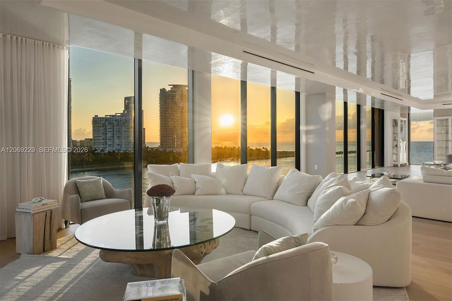 This sophisticated living room features a large, curved white sectional sofa arranged around a circular glass-topped coffee table with a natural wood base. The space is defined by expansive floor-to-ceiling windows that offer a panoramic view of the ocean and city skyline at sunset. The interior design is minimalist and elegant, complemented by light wood flooring and a glossy, reflective ceiling that enhances the airy, open atmosphere.