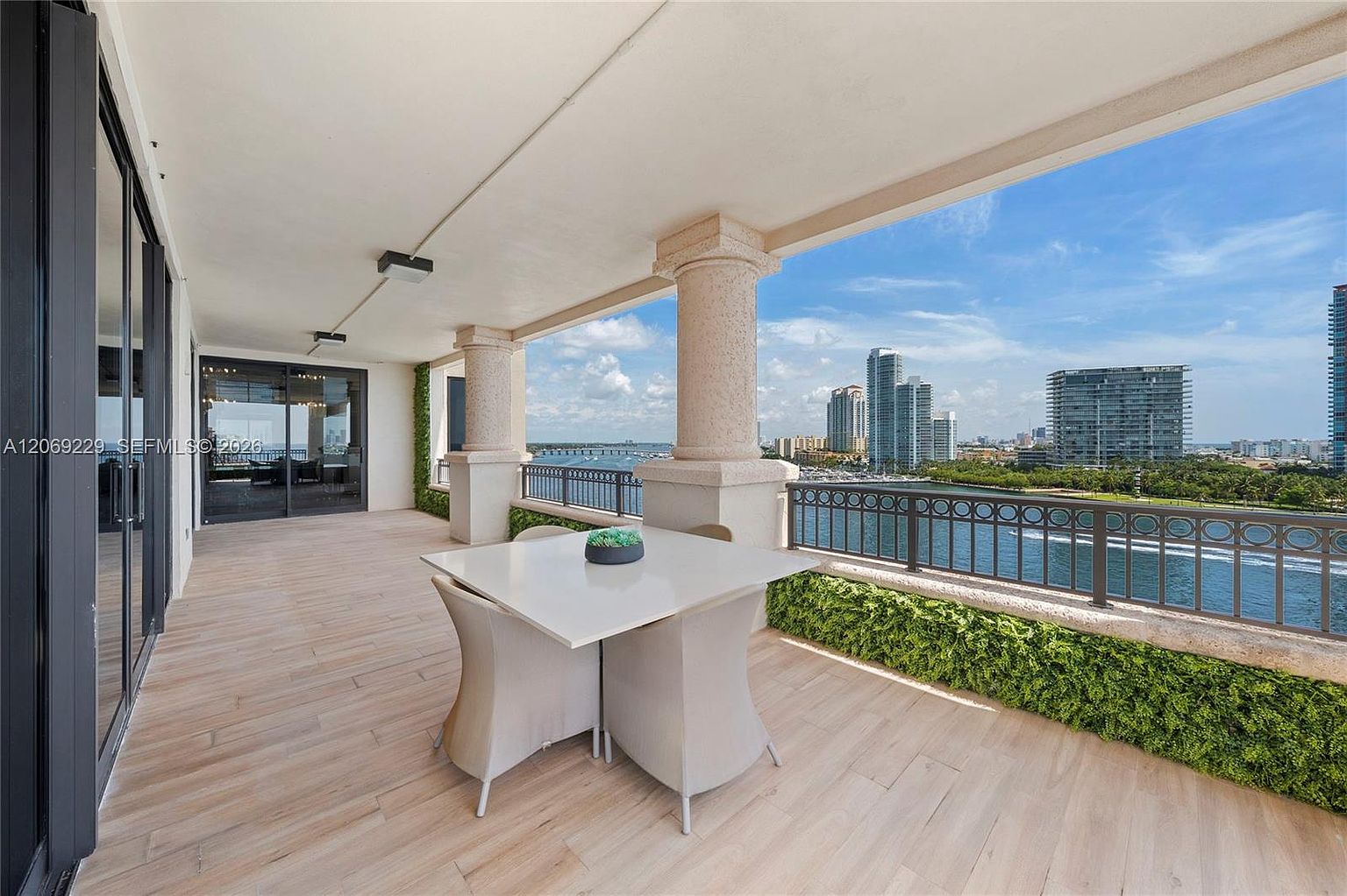 This expansive, covered balcony offers a sophisticated outdoor living space with light-toned wood-look flooring and a dining set positioned to enjoy the panoramic water and city skyline views. The area is framed by sturdy columns and a metal railing, with integrated greenery adding a touch of nature to the urban setting. The perspective is wide and inviting, showcasing the seamless transition between the indoor living area and the scenic outdoor terrace.