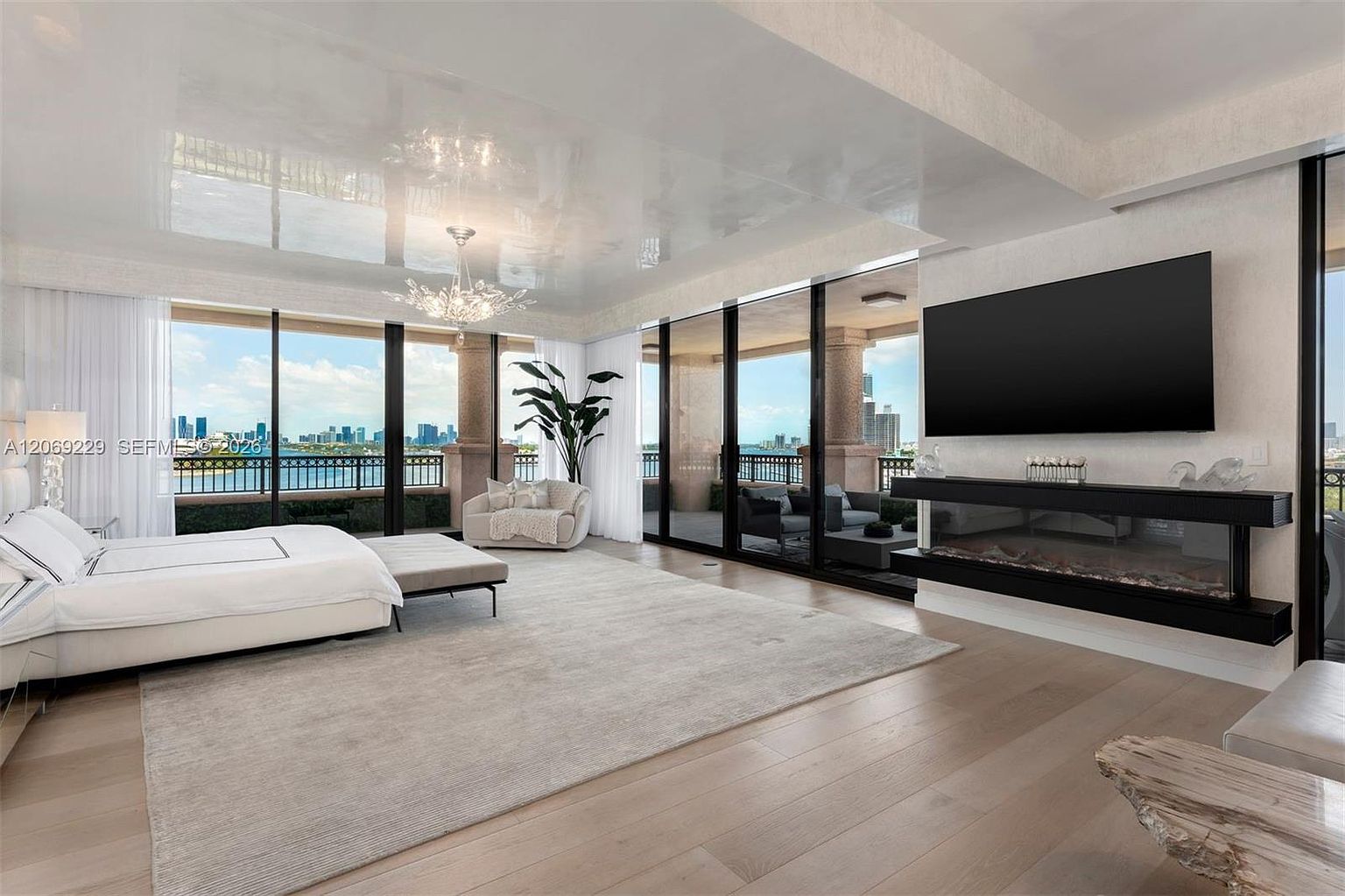 This expansive primary bedroom features a high-gloss reflective ceiling and light wood flooring, creating a bright and airy atmosphere. A large bed sits on a neutral area rug, positioned to face a sleek, linear fireplace integrated into a dark, horizontal wall feature topped by a large wall-mounted television. The room is framed by expansive glass walls that provide panoramic city and water views, leading out to a spacious private balcony.