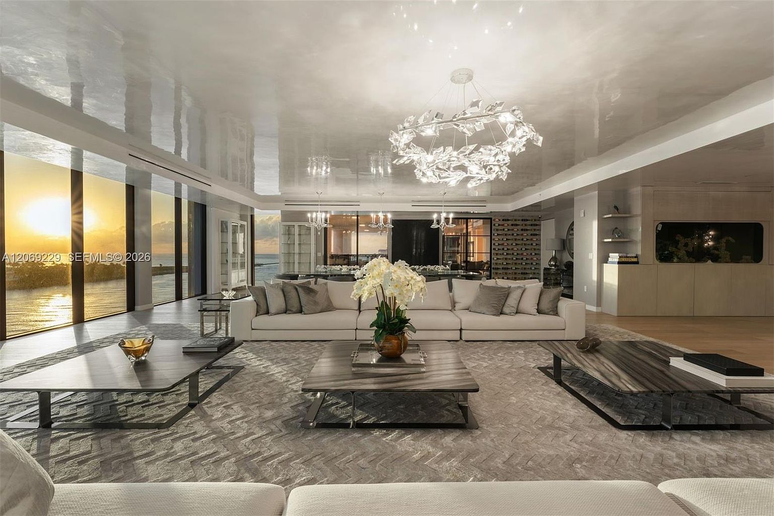 This expansive, modern living room features a large, neutral-toned sectional sofa arranged around three low-profile, dark-toned coffee tables. The space is defined by a high-gloss, reflective ceiling and floor-to-ceiling windows that provide a panoramic view of the sunset over the water. A striking, sculptural chandelier serves as a focal point, while a textured area rug adds depth to the open-concept layout.