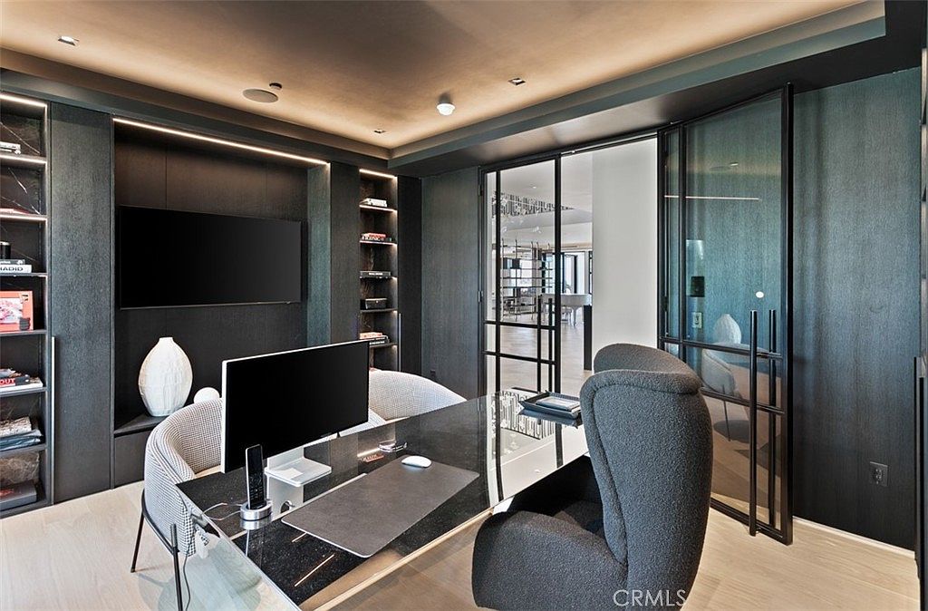 This sophisticated home office features a sleek, dark-toned aesthetic with built-in shelving, a modern glass-topped desk, and a high-backed ergonomic chair. The room is framed by elegant black-framed glass doors that provide a view into an adjacent living space, creating a seamless and open atmosphere. The combination of recessed lighting and minimalist decor gives the space a professional, high-end, and contemporary feel.