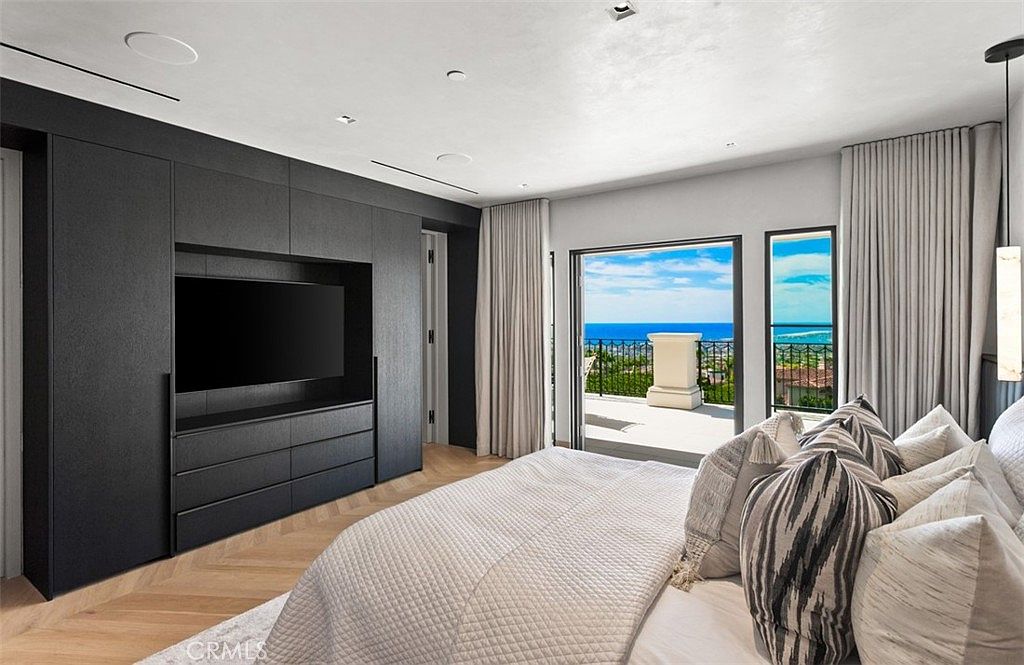 This luxurious primary bedroom features a sophisticated dark charcoal built-in media wall that contrasts beautifully with the light herringbone wood flooring. A large sliding glass door opens onto a private balcony, offering breathtaking, unobstructed views of the ocean and coastline. The room is elegantly furnished with a plush, textured bed and soft, neutral-toned curtains, creating a serene and high-end atmosphere.