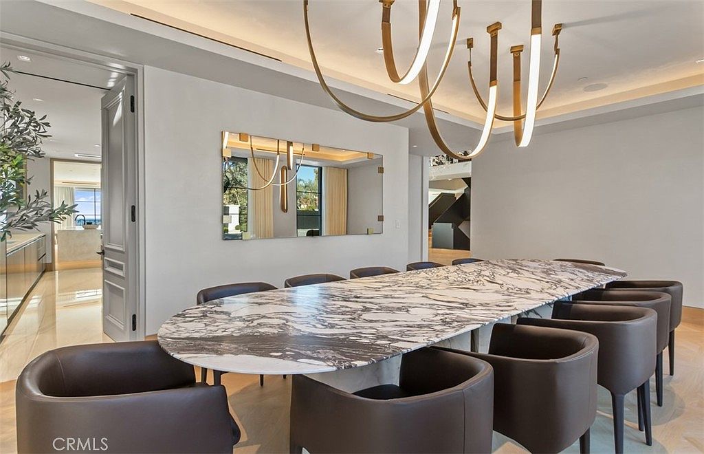 This elegant dining room features a long, oval-shaped marble table with dramatic black and white veining, surrounded by sleek, dark leather-upholstered chairs. A large, modern gold-toned chandelier hangs above the table, while a rectangular mirror on the wall reflects the space and adds depth. The room is finished with light-colored walls and flooring, creating a sophisticated and airy atmosphere that flows into an adjacent kitchen area.