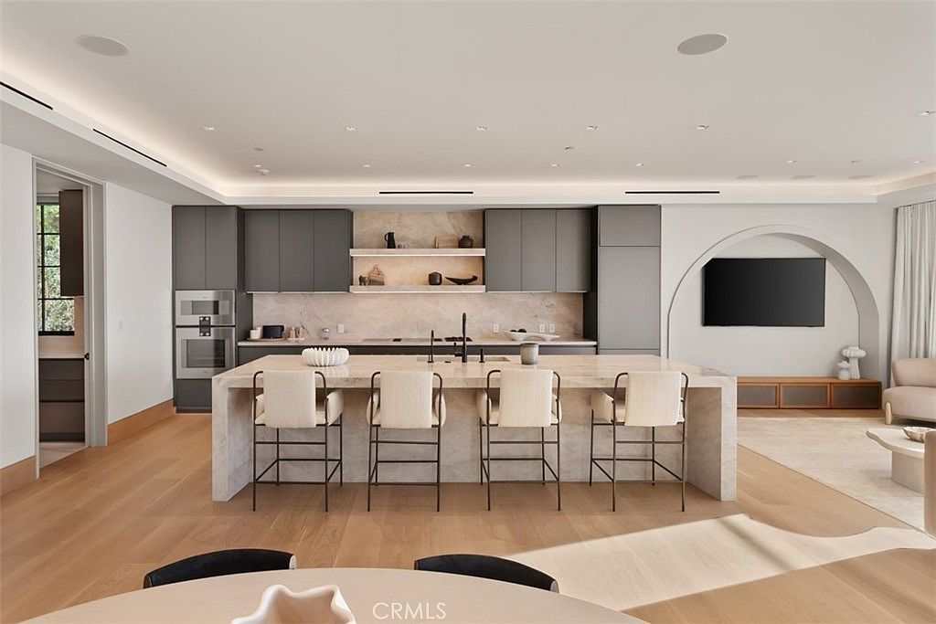 This sophisticated, modern kitchen features a large, light-toned stone island with four upholstered bar stools, serving as the centerpiece of the open-concept space. The cabinetry is a sleek, matte charcoal gray, complemented by warm wood flooring and integrated lighting that highlights the textured stone backsplash and floating shelves. The perspective is a wide, eye-level shot that captures the seamless flow between the kitchen and the adjacent living area, which features an elegant arched wall niche.