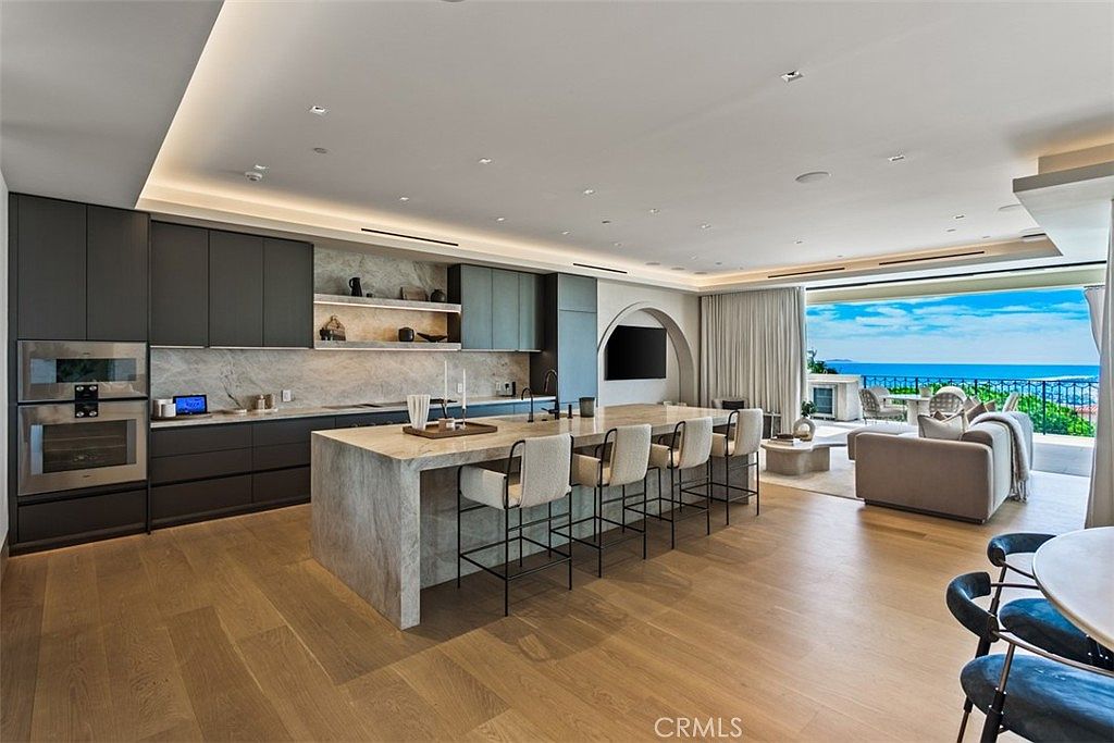 This expansive, modern open-concept kitchen features sleek dark cabinetry, a massive marble-topped island with bar seating, and high-end built-in appliances. The space flows seamlessly into a bright living area that opens up to a stunning panoramic ocean view through floor-to-ceiling glass doors. The design aesthetic is sophisticated and minimalist, emphasizing natural light and a luxurious indoor-outdoor lifestyle.
