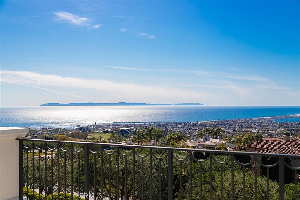This image captures a breathtaking, elevated view from a balcony, showcasing a panoramic vista of the ocean and a distant island under a clear blue sky. In the foreground, a decorative black wrought-iron railing frames the scene, overlooking a lush, sun-drenched coastal landscape with residential homes nestled among trees. The perspective offers a serene and luxurious atmosphere, emphasizing the property's prime location and stunning natural scenery.