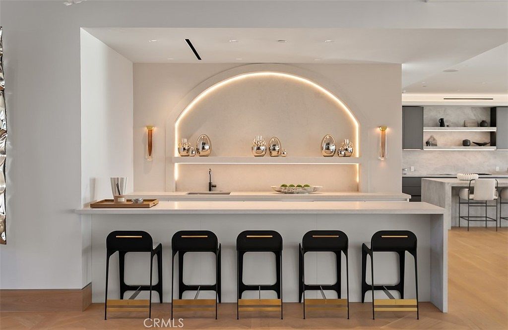 This sophisticated, modern kitchen features a striking arched alcove with integrated LED lighting that serves as a focal point above the sink. A long, minimalist island with a waterfall-edge countertop provides ample seating with five sleek, black bar stools. The neutral color palette and clean lines create an elegant, high-end atmosphere perfect for contemporary living.