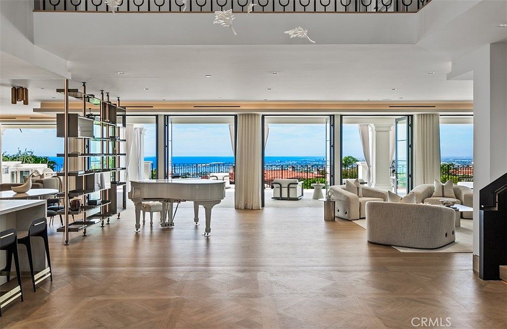 This expansive, open-concept living area features light-toned hardwood flooring and a sophisticated, neutral color palette. A grand white piano serves as a focal point, while floor-to-ceiling glass doors open to a stunning panoramic ocean view. The space is elegantly furnished with plush, contemporary seating, creating a luxurious and airy atmosphere perfect for high-end entertaining.