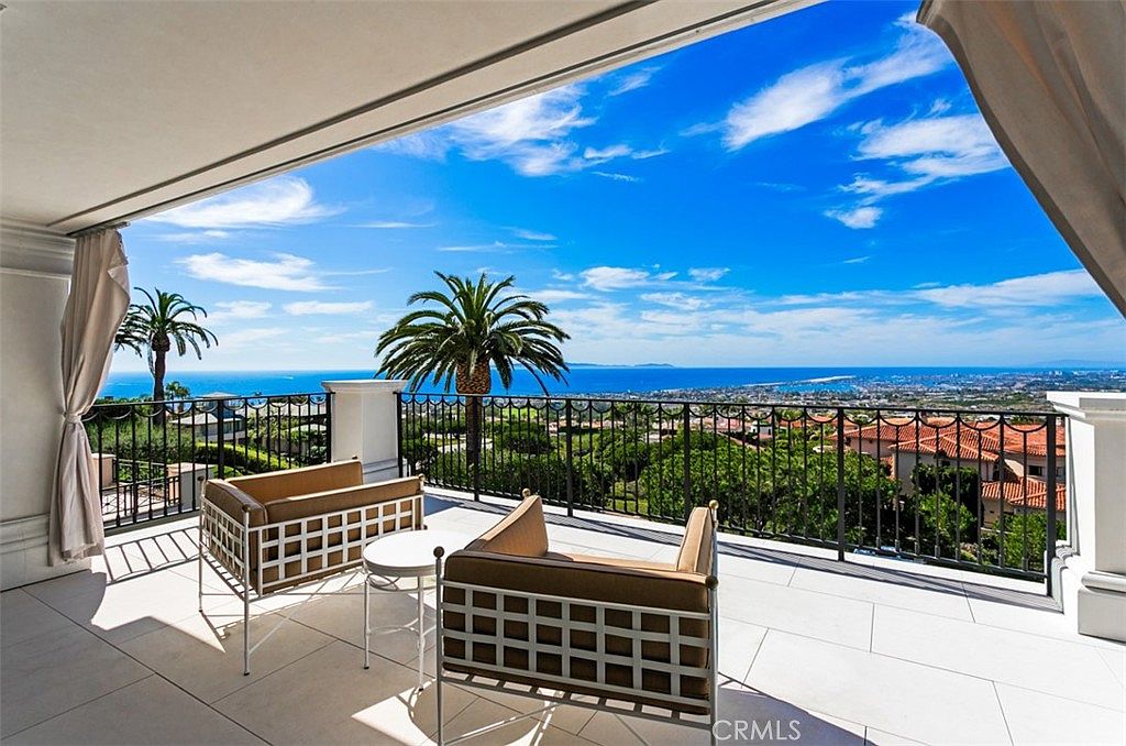 This luxurious covered balcony offers a breathtaking panoramic view of the ocean and coastline, framed by elegant architectural columns and sheer curtains. The space is furnished with two modern, cushioned armchairs and a small side table, creating a serene outdoor lounge area. The bright, sunny atmosphere and expansive horizon view highlight the premium nature of this property's outdoor living space.