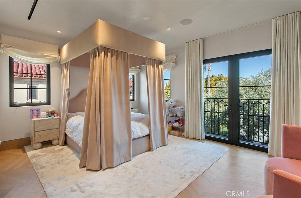 This elegant bedroom features a prominent canopy bed draped in soft, neutral-toned curtains, serving as the room's focal point. The space is bright and airy, with large French doors leading to a balcony and light-colored hardwood flooring throughout. A plush pink armchair and a matching nightstand add a touch of warmth and sophistication to the serene, well-lit interior.